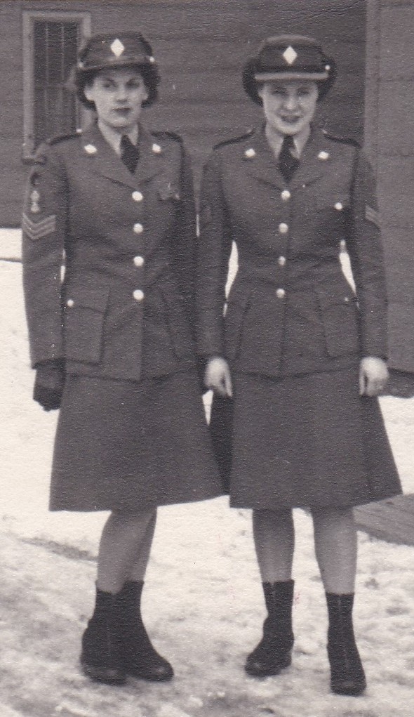 Deux femmes habillées en uniformes identiques sont debout devant un bâtiment. Les uniformes consistent d’une veste avec des boutons en laiton et une jupe à l’auteur du genou. Les femmes portent aussi des bottes. Elles ont des cheveux courts et elles portent un bonnet de l’armée.