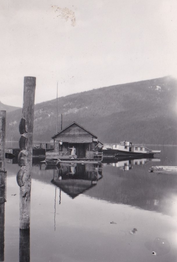 Une femme est debout dans une maison en bois qui flotte sur l’eau. Il y a des grosses montagnes autour de l’eau. Deux autres vaisseaux sont attachés à la maison en bois.