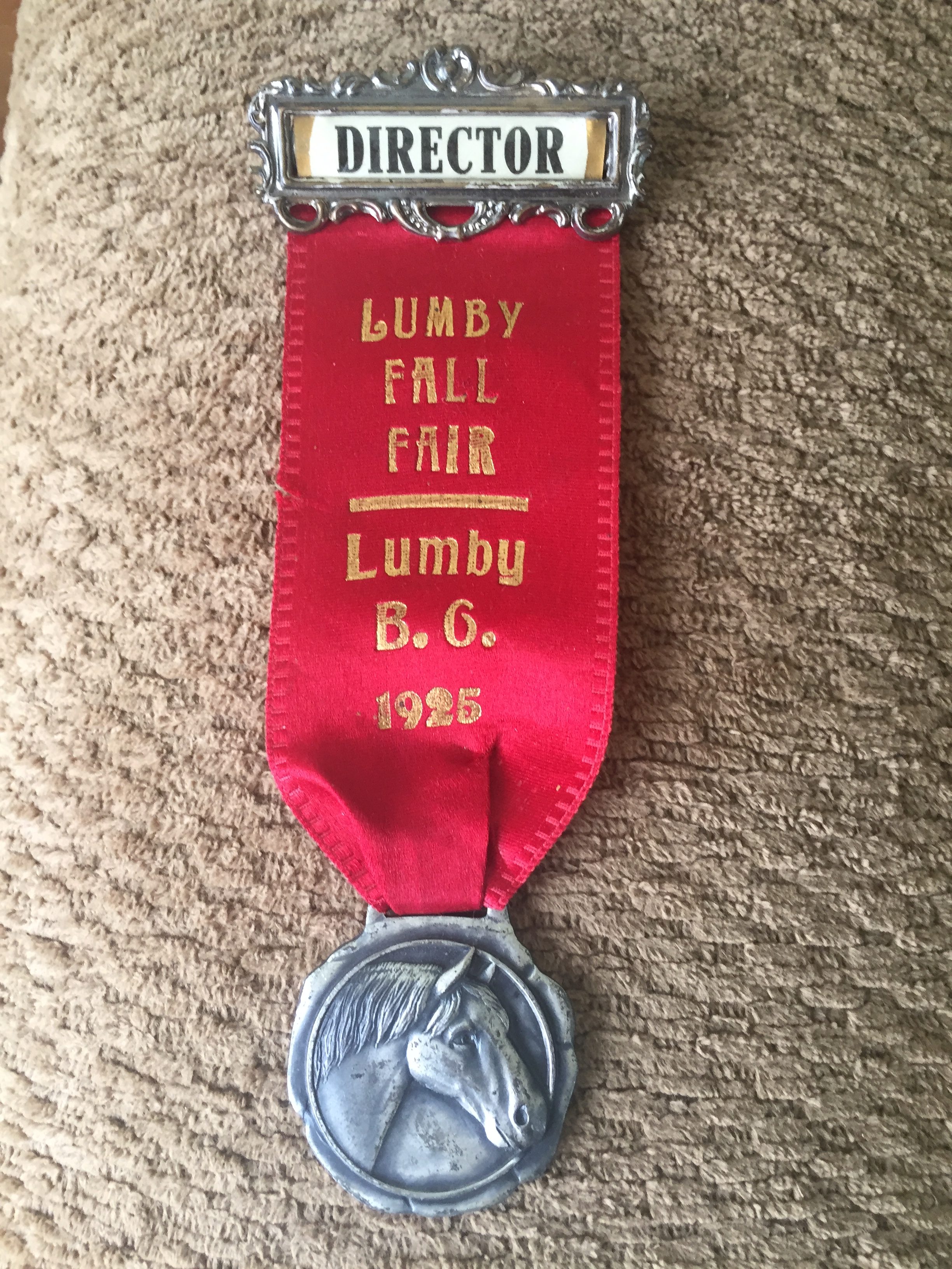 Le directeur porte un badge avec un ruban rouge où il est écrit : « La foire d’automne de Lumby C.B. 1925 ». Il y a un morceau de métal avec la tête d’un cheval attaché au ruban.