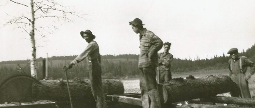 Quatre hommes travaillent à côté d’une scierie rudimentaire. La scie coupe une bûche de mélèze. Une autre bûche est sur un chariot pour être coupée après. La scierie est au milieu d’un champ.