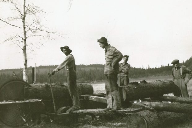 Quatre hommes travaillent à côté d’une scierie rudimentaire. La scie coupe une bûche de mélèze. Une autre bûche est sur un chariot pour être coupée après. La scierie est au milieu d’un champ.