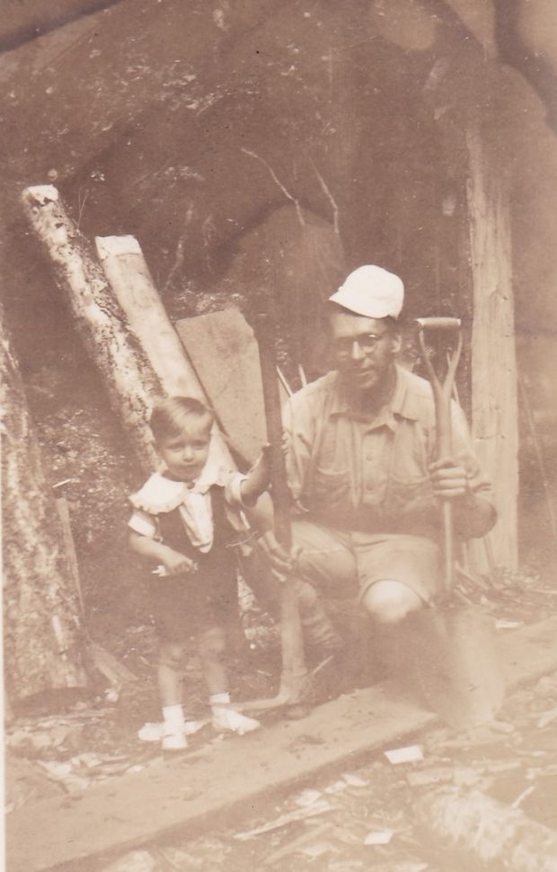 Un petit garçon est debout à côté de son père qui est à genou devant l’entrée d’une mine. L’homme tient une pelle et une pioche. L’homme porte le chapeau du petit garçon.