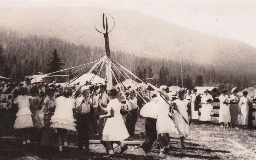 Un groupe d’enfants danse autour de l’arbre de mai. Les filles portent des robes froncées et les garçons portent des pantalons noirs et des chemises. Un groupe de pionniers les regardent.