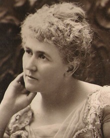 Photo portrait d’une femme légèrement tournée vers la droite, avec le bout de la main droite touchant son visage. Ses cheveux sont ramassés en chignon. 
