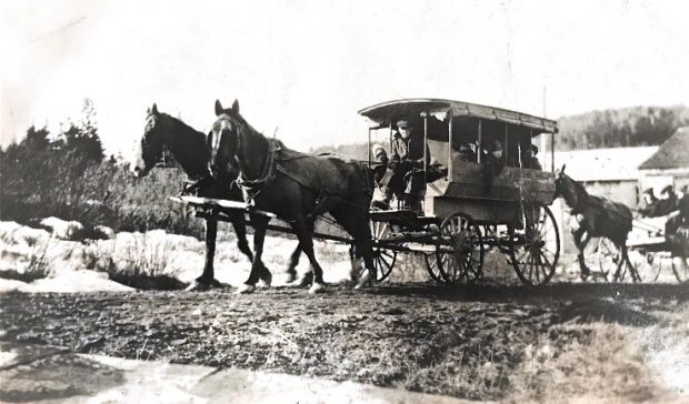 Vue de gauche de fourgons scolaires tirés par des chevaux, avec des conducteurs et des enfants habillés en vêtements d’hiver, descendant une route de terre bordée de neige de chaque côté. Le premier fourgon est couvert et tiré par deux chevaux, le second est découvert et tiré par un seul cheval. 