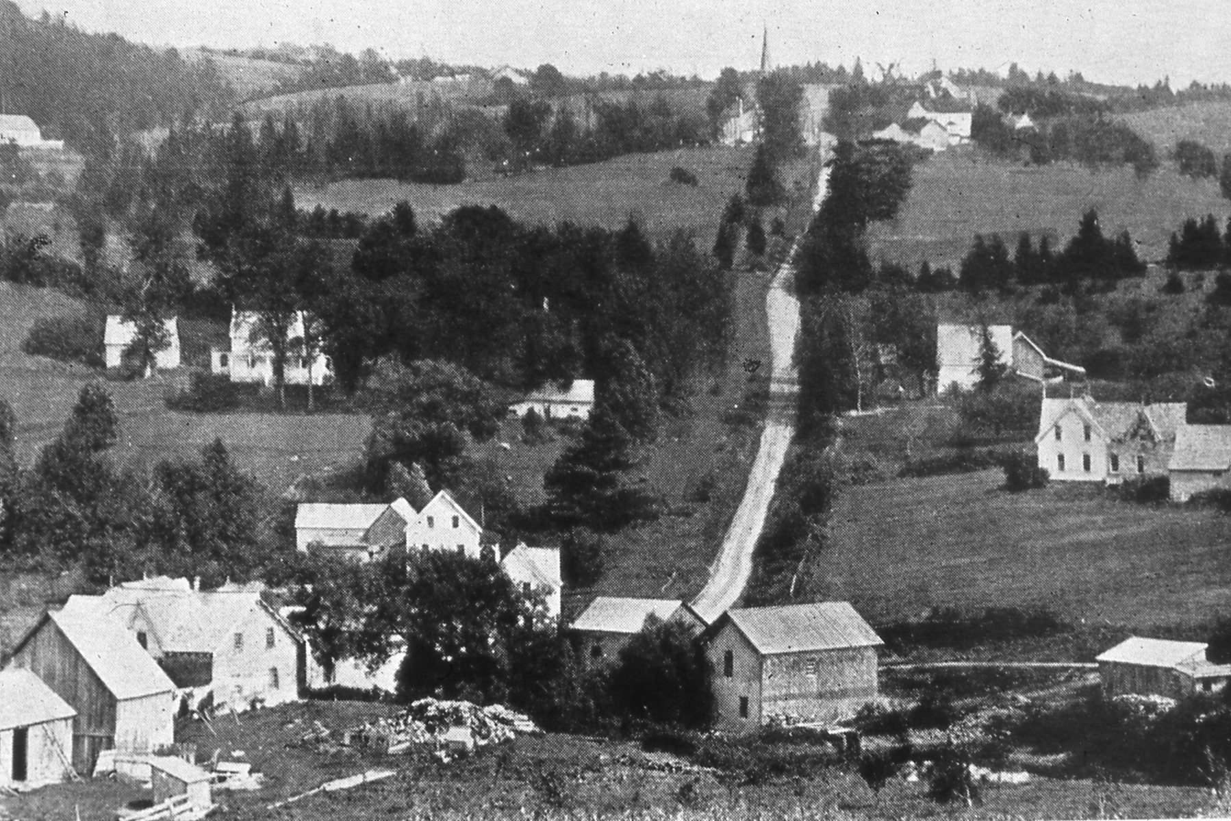 Vers 1900. Paysage rural montrant une église au loin, au sommet d’une colline. Des maisons et des fermes apparaissent de part et d’autre de la colline qui descend vers la partie basse du village. 