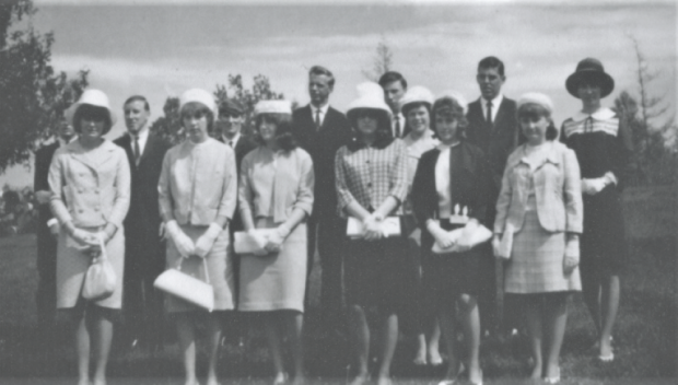 Groupe de diplômés comprenant huit jeunes femmes portant des tailleurs à jupe et des chapeaux. Six jeunes hommes en costume sont debout derrière elles. 