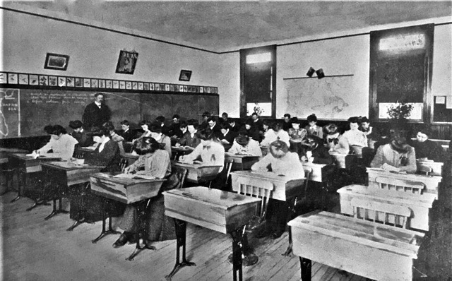 Un enseignant, dos au tableau, surveille une trentaine d’élèves assis à leur bureau en train de passer un examen. 