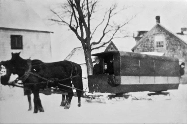 Vue de gauche d’un fourgon scolaire couvert sur patins, tiré par deux chevaux quittant une ferme par une journée d’hiver. 