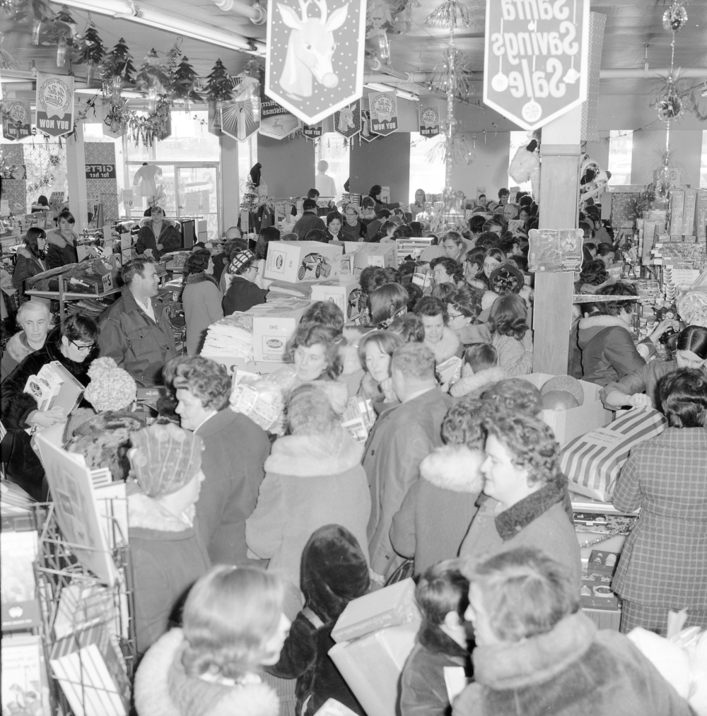 Photographie d'archives en noir et blanc. Plus de 70 personnes en manteaux d’hiver et chapeaux font la file chez Cohen. Des décorations de Noël et des bannières pendent du plafond.