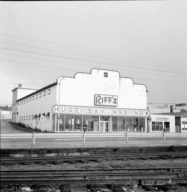 Photographie d'archives en noir et blanc. Vue de la rue. Un bâtiment est visible de l’autre côté des voies de chemin de fer. Texte du panneau accroché en travers du bâtiment : RIFF’S. En dessous : HUGE SAVINGS NOW (GROS RABAIS MAINTENANT). On aperçoit des mannequins hommes et femmes à travers les fenêtres de l’avant du bâtiment. Le magasin P.F. Kearney est à droite.