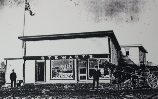 Photographie d'archives en noir et blanc. Vue de la rue. Le bâtiment principal a deux portes d’entrée et deux grandes fenêtres. Un Union Jack flotte à gauche au-dessus du bâtiment. Deux hommes sont debout dehors et un autre homme conduit une charrette à cheval.