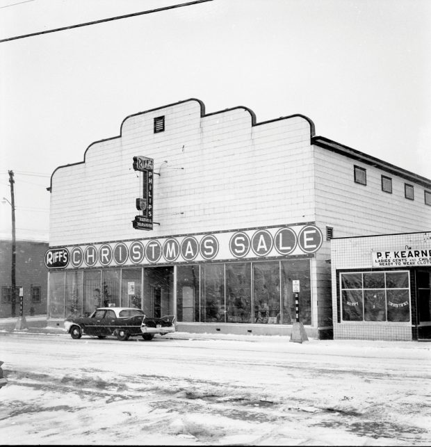 Photographie d'archives en noir et blanc. Vue de la rue. Vue extérieure des magasins Riff’s et P.F. Kearney. On peut distinguer le décor de Noël dans les fenêtres des deux magasins. La neige tombe et une voiture est garée en face de Riff’s. Le panneau au-dessus du magasin Riff’s indique : RIFF’S CHRISTMAS SALE (SOLDES DE NOËL DE RIFF’S).