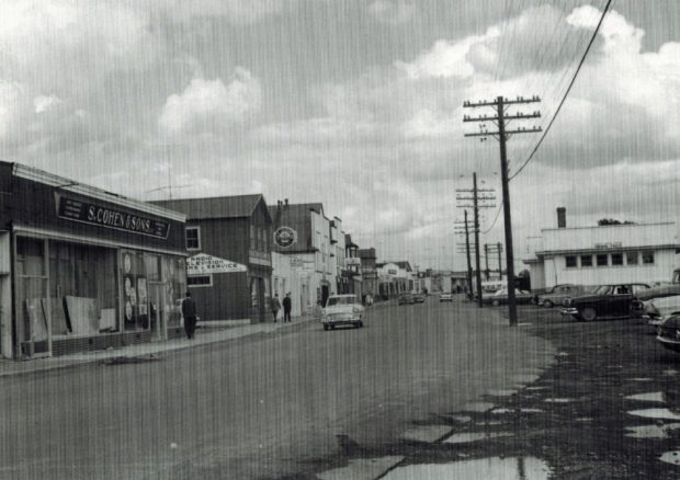 Photographie d'archives en noir et blanc. Vue de la rue. À gauche de la photo, des commerces de la rue Main. À droite, des poteaux téléphoniques et la gare ferroviaire. Il y a quatre voitures dans la rue et plusieurs piétons sur le trottoir, à gauche de la photo.