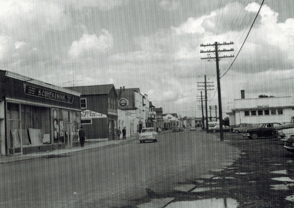 Photographie d'archives en noir et blanc. Vue de la rue. À gauche de la photo, des commerces de la rue Main. À droite, des poteaux téléphoniques et la gare ferroviaire. Il y a quatre voitures dans la rue et plusieurs piétons sur le trottoir, à gauche de la photo.