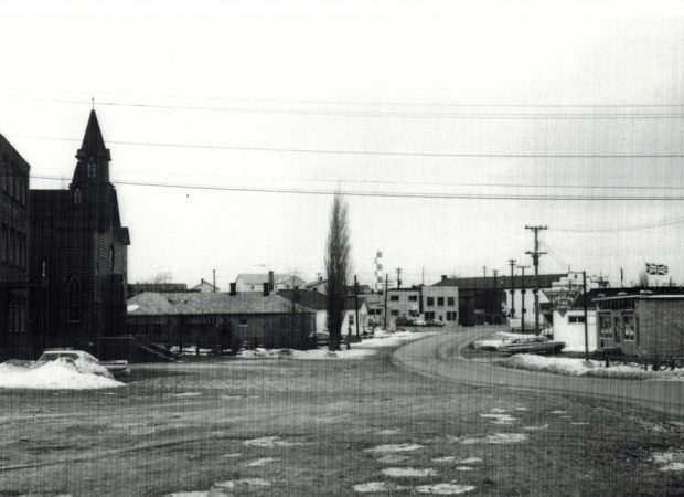 Photographie d'archives en noir et blanc. Vue de la rue. Vue vers le nord en direction de la rue Main de Windsor depuis l’église St Joseph, qui se trouve à gauche de la photo. À droite, on voit le bâtiment de Canadian Tire et, au centre, le panneau de signalisation des voies de chemin de fer.