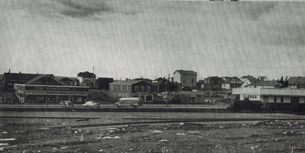 Photographie d'archives en noir et blanc. Vue de la rue. Le chemin de fer au premier plan et magasins en arrière-plan. À gauche on voit le magasin S. Cohen & Fils, à droite le train et la gare du chemin de fer de Terre-Neuve.