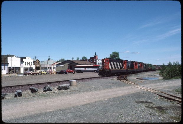 Photographie couleur. Vue en direction de la rue Main depuis l’autre côté des voies de chemin de fer. À droite, on voit le train CN et au centre, la gare ferroviaire.