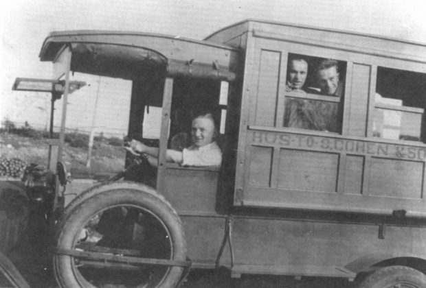 Photographie d'archives en noir et blanc. Un homme conduit un bus qui compte deux passagers à la première fenêtre. Sur le côté du bus est écrit : BUS TO S. COHEN & SONS (BUS VERS S. COHEN & FILS).