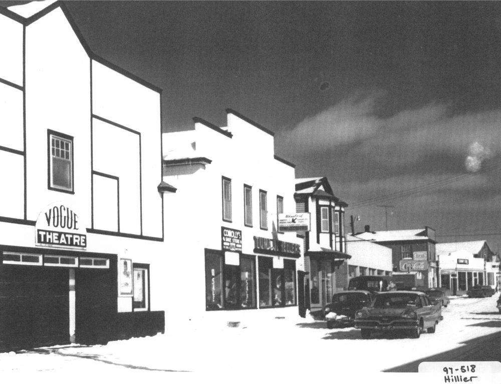 Photographie d'archives en noir et blanc. Vue de la rue. Au centre de la photo et à droite du cinéma Vogue se trouvent le magasin de chaussures Connolly’s et la bijouterie Tuma.