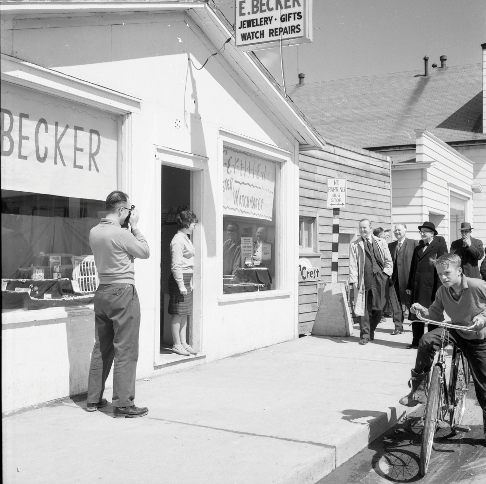 Photographie d'archives en noir et blanc. Vue de la rue. Vue extérieure de la bijouterie d’E. Becker. Un homme à gauche de la photo prend une photo, une femme se tient sur le seuil du magasin, un groupe de six hommes en costume marche en direction du magasin et un garçon passe à vélo.