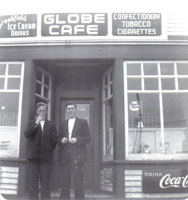 Photographie noir et blanc. Deux hommes debout sur les marches du Café Globe, entre les deux fenêtres en baie. Textes des panneaux situés au sommet de l’immeuble, de gauche à droite : Brookfield ICE CREAM DRINKS, GLOBE CAFE, et CONFECTIONERY, TOBACCO, et CIGARETTES (CRÈMES GLACÉES Brookfield, BOISSONS, CAFÉ GLOBE, et CONFISERIE, TABAC, et CIGARETTES). Texte du panneau en bas à droite : DRINK Coca-Cola (BUVEZ Coca-Cola).
