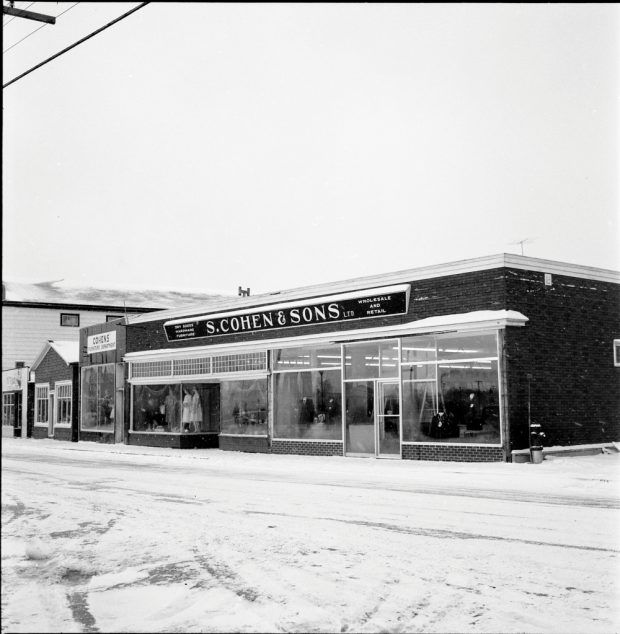 Photographie d'archives en noir et blanc. Vue de la rue. Vue des magasins S. Cohen & Sons et Cohen’s Furniture Department, en direction de l’ouest de la rue Main. Au-dessus du magasin on peut lire : DRY GOODS; HARDWARE; FURNITURE; S. COHEN & SONS LTD.; WHOLESALE AND RETAIL (MERCERIE ; QUINCAILLERIE ; MEUBLES ; S. COHEN & FILS LTD ; VENTE EN GROS ET AU DÉTAIL). Des mannequins homme et femme sont visibles dans les vitrines du magasin.