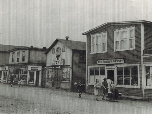 Photographie d'archives en noir et blanc. Vue de la rue. Rue Main de Windsor en direction de l’ouest. On voit le Brown Derby, le cinéma King Edward, les magasins A. Peckford et S. Cohen & Fils et le café Purity. Kirk Pomeroy porte un balai en bouleau, Daisy Bennett porte un manteau léger et Mme Pomeroy pousse une poussette.