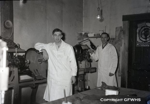 Photographie d'archives en noir et blanc. Deux hommes en blouses de laboratoire sont debout chez le cordonnier Hermann Münch.