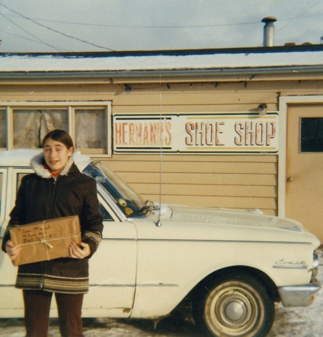 Photographie couleur. Vue de la rue. Une fillette portant un manteau d’hiver et tenant un paquet se tient devant une voiture blanche, devant la boutique du cordonnier Hermann’s Shoe Shop.