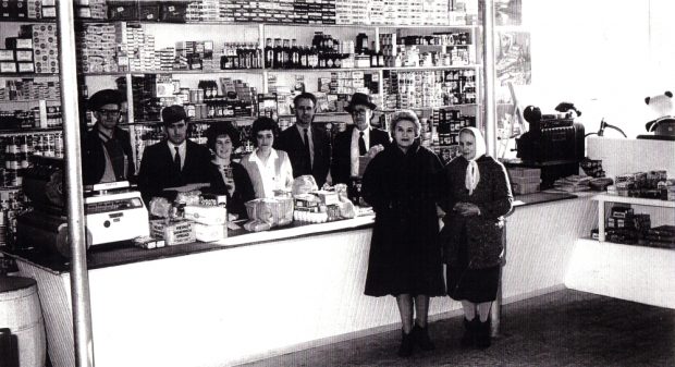 Photographie d'archives en noir et blanc. Quatre hommes et deux femmes se tiennent derrière la caisse et le comptoir, et deux femmes en manteau devant le comptoir. Les étagères situées derrière la caisse sont remplies d’emballages, de boîtes de conserve et de bouteilles de produits.