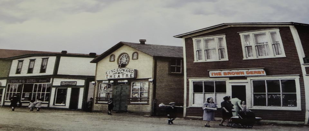 Photographie d'archives couleur. Vue de la rue Main de Windsor en direction de l’ouest. On voit le Brown Derby, le cinéma King Edward, les magasins A. Peckford et S. Cohen & Fils et le café Purity. Kirk Pomeroy porte un balai en bouleau, Daisy Bennett porte un manteau bleu-gris et Mme Pomeroy pousse une poussette.