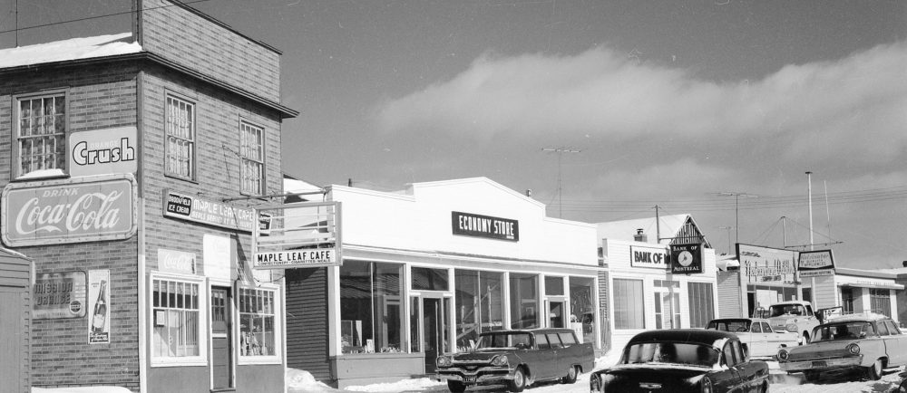 Photographie d'archives en noir et blanc. Vue de la rue. Vue de la rue Main de Windsor en direction de l’est. On voit le café Maple Leaf, le magasin Economy Store, la Banque de Montréal, le magasin Newfoundland Outfitting et le bureau de poste de Windsor. Plusieurs voitures sont dans la rue, qui est enneigée.