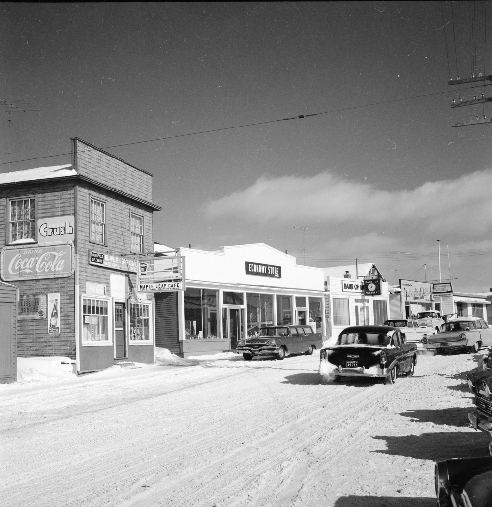 Photographie d'archives en noir et blanc. Vue de la rue. Vue de la rue Main de Windsor en direction de l’est. On voit le café Maple Leaf, le magasin Economy Store, la Banque de Montréal, le magasin Newfoundland Outfitting et le bureau de poste de Windsor. Plusieurs voitures sont dans la rue, qui est enneigée.