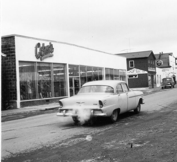 Photographie d'archives en noir et blanc. Vue de la rue. Magasin Cohen’s, sur la rue Main en direction de l’est vers Great Eastern Oil, et magasin de E. Becker. Des mannequins femmes sont visibles à gauche des portes principales du magasin Cohen’s. Voiture au centre de l’image.