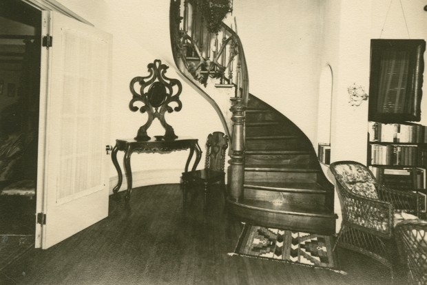 Photo noir et blanc. Hall d'entrée avec un grand escalier de bois au centre.