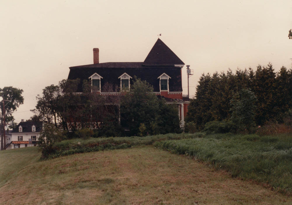 Photo couleur. Au centre, une maison à l’abandon. À l'avant plan, un terrain non entretenu.
