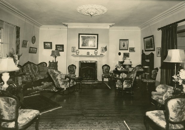 Photo noir et blanc. Salon au décor surchargé. Des meubles sont disposés de chaque côté de la pièce avec au centre, un foyer.