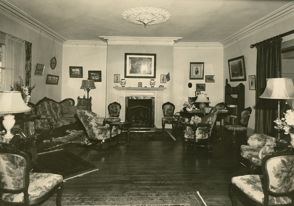 Photo noir et blanc. Salon au décor surchargé. Des meubles sont disposés de chaque côté de la pièce avec au centre, un foyer.