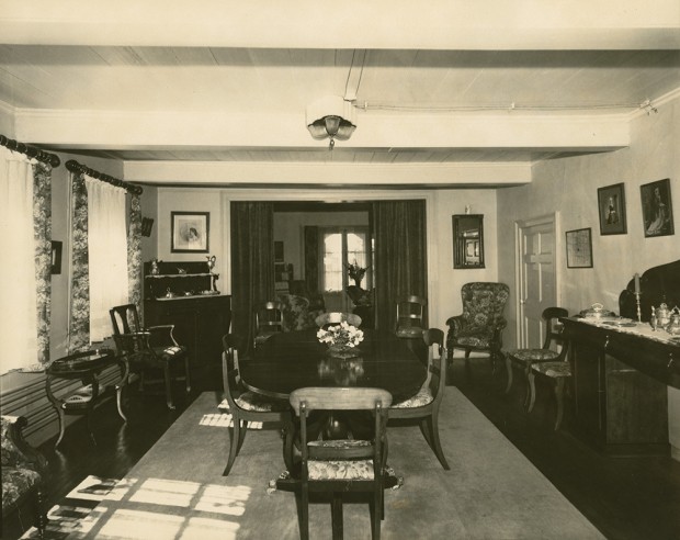 Photo noir et blanc. Salle à manger. La table est disposée au centre, sur un grand tapis. À gauche, un immense buffet est adossé au mur. On devine le salon en arrière-plan.