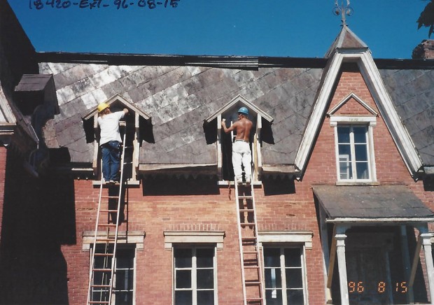 Photo couleur. Ouvriers dans des échelles qui réparent les lucarnes de la façade du Manoir.