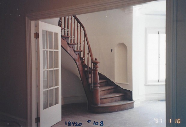 Photo couleur. Hall d'entrée vide avec l'escalier de bois au centre.