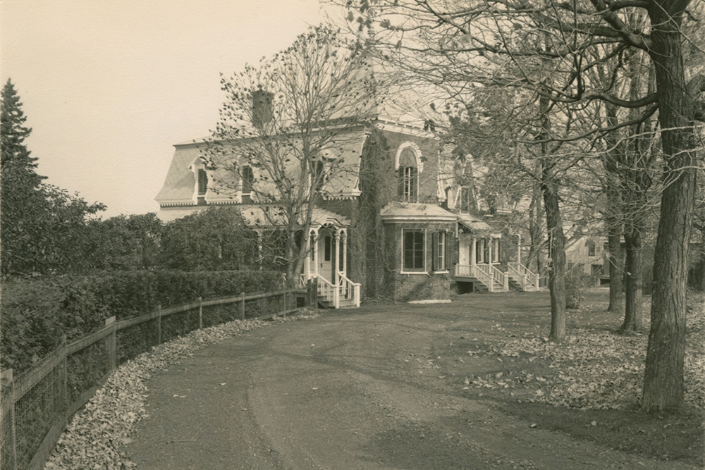 Photo noir et blanc. Un chemin à l'avant-plan mène à une maison entourée d'arbres en arrière-plan.