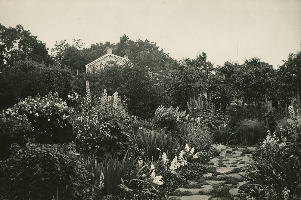 Photo noir et blanc. Jardin abondamment fleuri avec allée de pierre à l’avant-plan. De nombreux arbres matures cachent une maison à l’arrière-plan. 