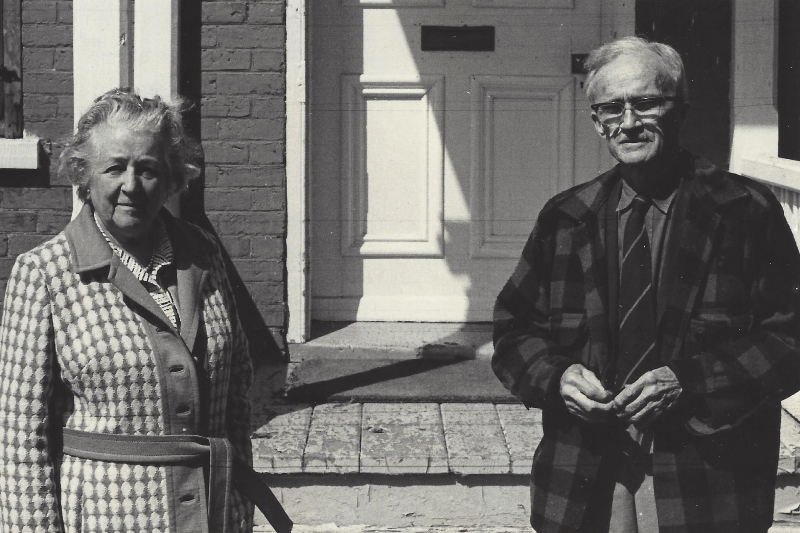 Photo noir et blanc. Deux personnes, une femme à gauche et un homme à droite prennent la pose à l’extérieur. À l’arrière, on devine la façade du Manoir Fraser.