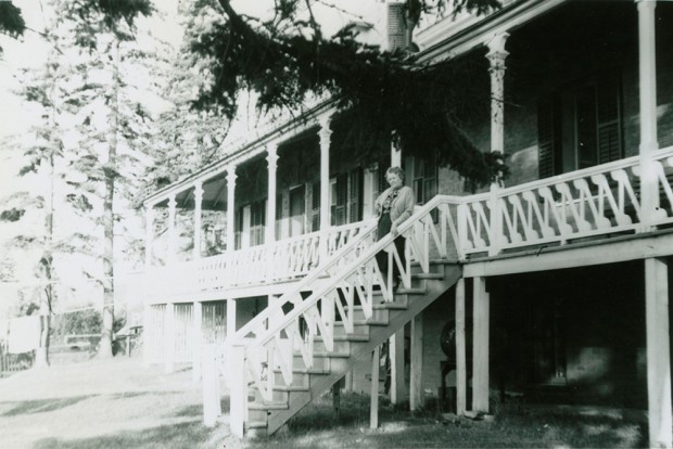 Photo noir et blanc. Femme âgée debout dans l’escalier extérieur qui donne sur la grande galerie entourant le manoir.