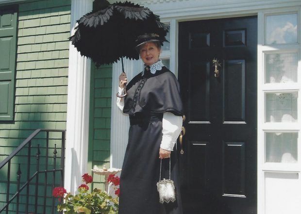 Photo couleur. Une femme, vêtue d’un costume d’époque prend la pose devant une maison de couleur verte. 