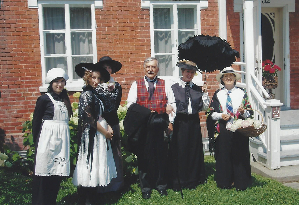 Photo couleur. Six personnes, cinq femmes et un homme, vêtus de costumes d’époque, prennent la pose devant une maison de briques. 