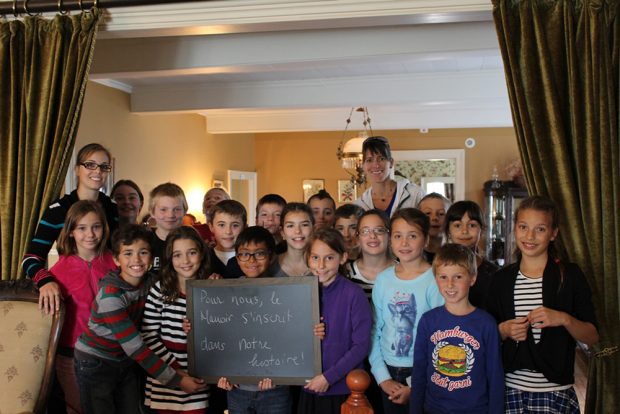 Photo couleur. Classe d'une vingtaine d'étudiants de niveau primaire prennent la pose. l'un d'eux tient un tableau noir sur lequel il est écrit ce que le manoir représente pour leur classe.