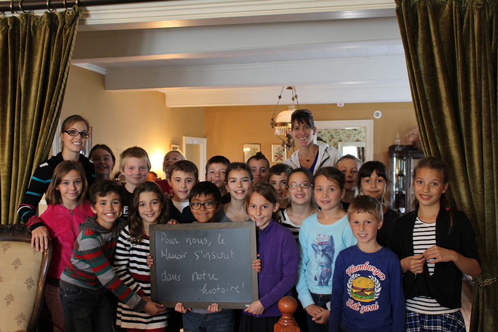 Photo couleur. Classe d'une vingtaine d'étudiants de niveau primaire prennent la pose. l'un d'eux tient un tableau noir sur lequel il est écrit ce que le manoir représente pour leur classe.